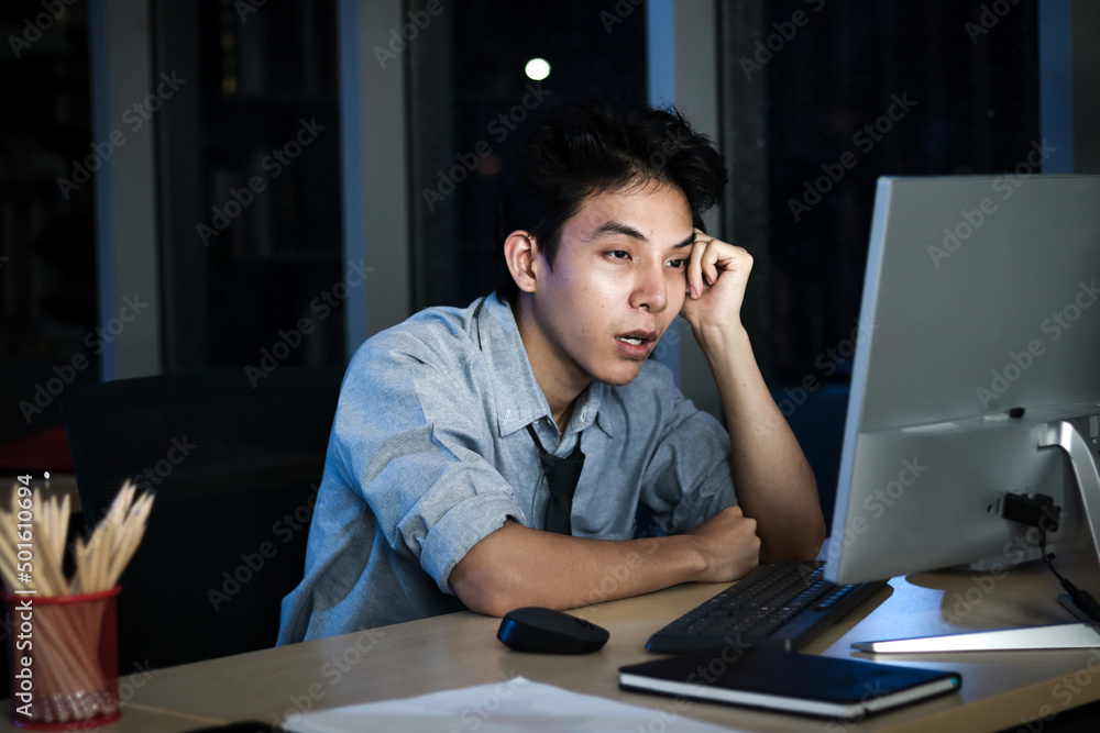 Asian young tired staff officer man using desktop computer having overwork project overnight in office, exhausted unhappy businessman feeling sleepy after after working hard overtime at night. Asian young tired staff officer man using desktop computer having overwork project overnight in office, exhausted unhappy businessman feeling sleepy after after working hard overtime at night.