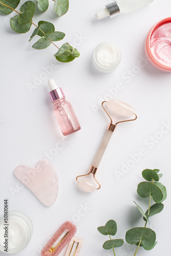 Skincare concept. Top view vertical photo of rose quartz roller gua sha pink eye patches glass bottles barrettes and eucalyptus on isolated white background