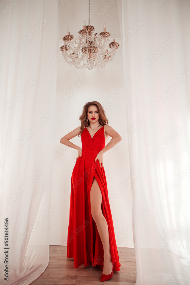 Fototapeta premium teen fashion. young blonde girl in long red dress is standing with hands near waist and looking straight with smile on the white wall background with crystal chandelier. fashion concept, free space