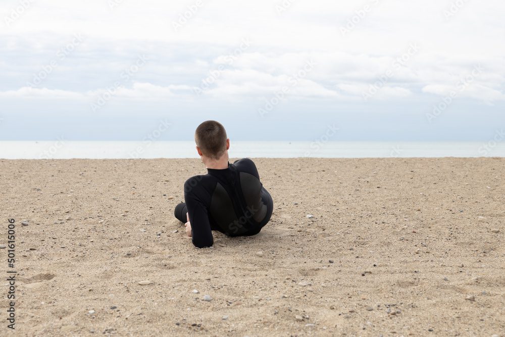 Man laying on one arm alone on a beach next to the ocean looking out wearing a black wetsuit before a training exercise on a cloudy overcast day on a lake. Man laying on one arm alone on a beach next to the ocean looking out wearing a black wetsuit before a training exercise on a cloudy overcast day on a lake.