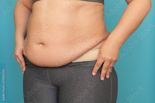 Fotografie Cropped photo of fat plump plus-size overweight woman wearing grey sports bra, showing excess naked stomach, taking off leggings with hands on blue background
