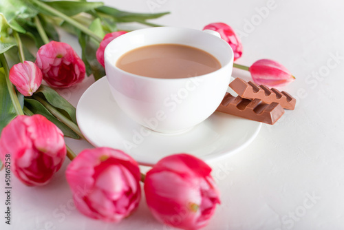Banner.One cup of hot morning coffee, milk chocolate and a bouquet of pink-lilac tulips on a bright white background. View from above. Copy space for text. Holidays concept.