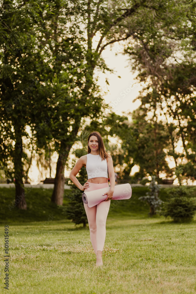 Brunette is standing on grass in park. Young smiling woman in white top and pink sweatpants is standing on green lawn and holding pink yoga and Pilates mat in her hands, posing and looking at camera. Brunette is standing on grass in park. Young smiling woman in white top and pink sweatpants is standing on green lawn and holding pink yoga and Pilates mat in her hands, posing and looking at camera.