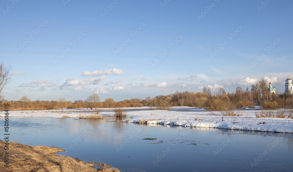 Fototapeta premium March sunny day by the river. A picturesque landscape, early spring, a river with snow-covered banks, dry grass and bushes. Church in the background. The first thaws, the snow is melting