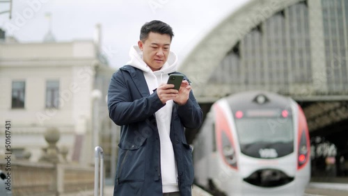 Asian tourist booking accommodation at home using mobile phone, passenger arrives by train to new city. Passengers at rairoad station and arriving train. Man use smart phone before travel photo