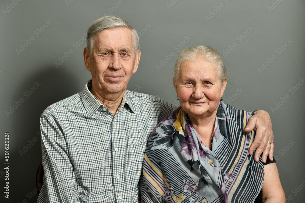 Studio portrait of an elderly couple on a gray background. The old man is hugging his wife, they are smiling, happy. Studio portrait of an elderly couple on a gray background. The old man is hugging his wife, they are smiling, happy.
