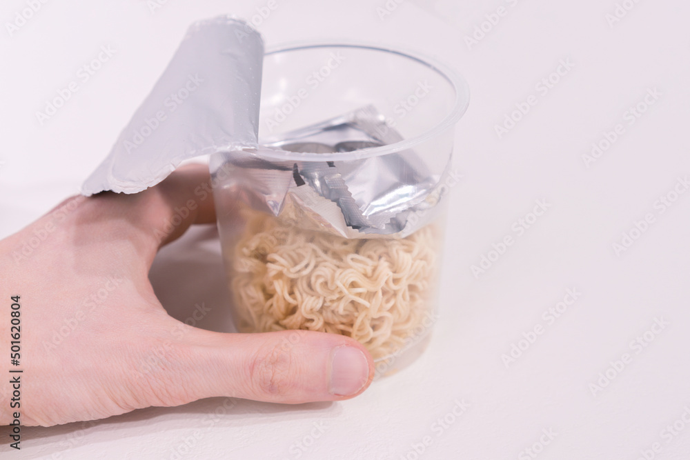 The man’s hand holds a transparent noodle cup with a package of