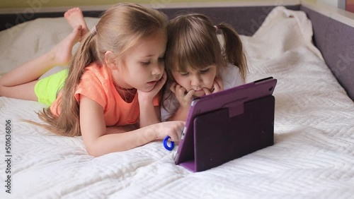 Two beautiful little sisters lying in the bed and look at the screen of a tablet, smart kids using smart technology