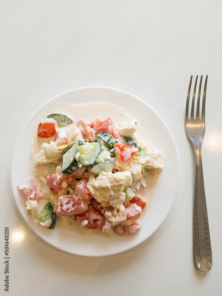 Delicious salad with tomatoes, cucumbers, Chinese cabbage, feta cheese and mushroom mayonnaise on a white plate and white background. A fork is placed next to it. Delicious salad with tomatoes, cucumbers, Chinese cabbage, feta cheese and mushroom mayonnaise on a white plate and white background. A fork is placed next to it.