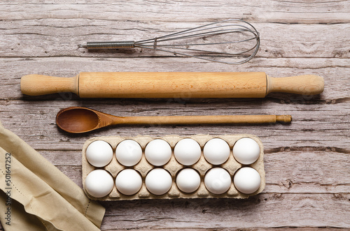 A whisk, a rolling pin, a wooden spoon, some white eggs in an egg container and a piece of cloth on a wooden table. 