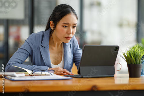 Confident Asian female business analyst financial advisor preparing statistic report studying documents on a work desk, browsing information online using a tablet, writing out notes on the paper sheet