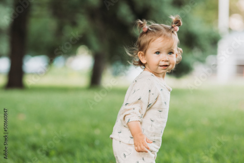 cute little smiling girl with a funny hairstyle hides behind trees and runs and plays in the park