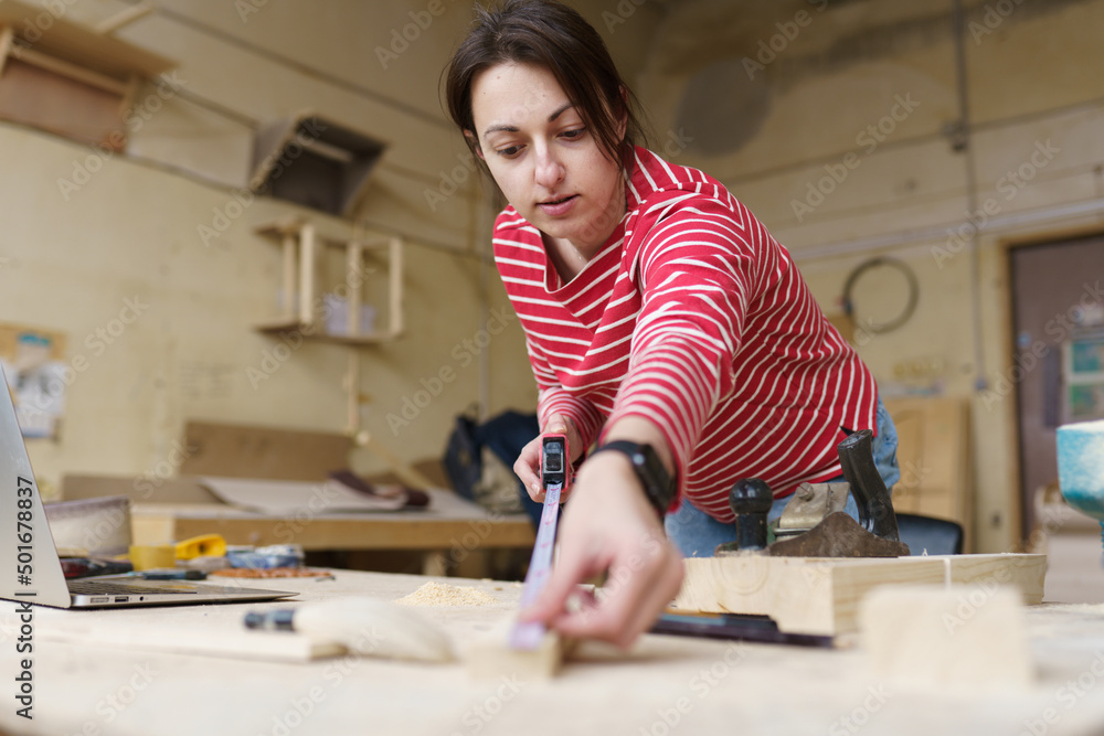 A female carpenter in a red striped shirt works with a ruler and makes marks on a tree at a table in the workshop