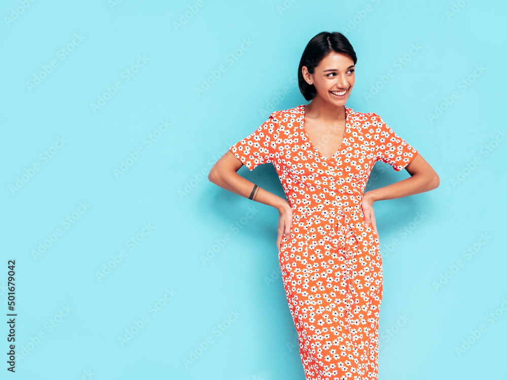 Obraz premium Young beautiful smiling female in trendy summer red dress. Sexy carefree woman posing near blue wall in studio. Positive model having fun. Cheerful and happy. Isolated