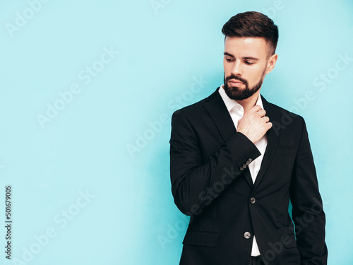 Portrait of handsome confident stylish hipster lambersexual model. Sexy modern man dressed in elegant black suit. Fashion male posing in studio near blue wall