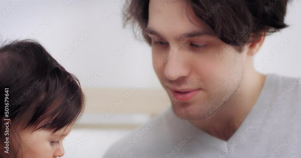 Close-up of a charming little girl with dark hair and brown eyes, whom dad feeds with a spoon with vegetable puree. The first meal of a child. Parents give their child healthy food Close-up of a charming little girl with dark hair and brown eyes, whom dad feeds with a spoon with vegetable puree. The first meal of a child. Parents give their child healthy food