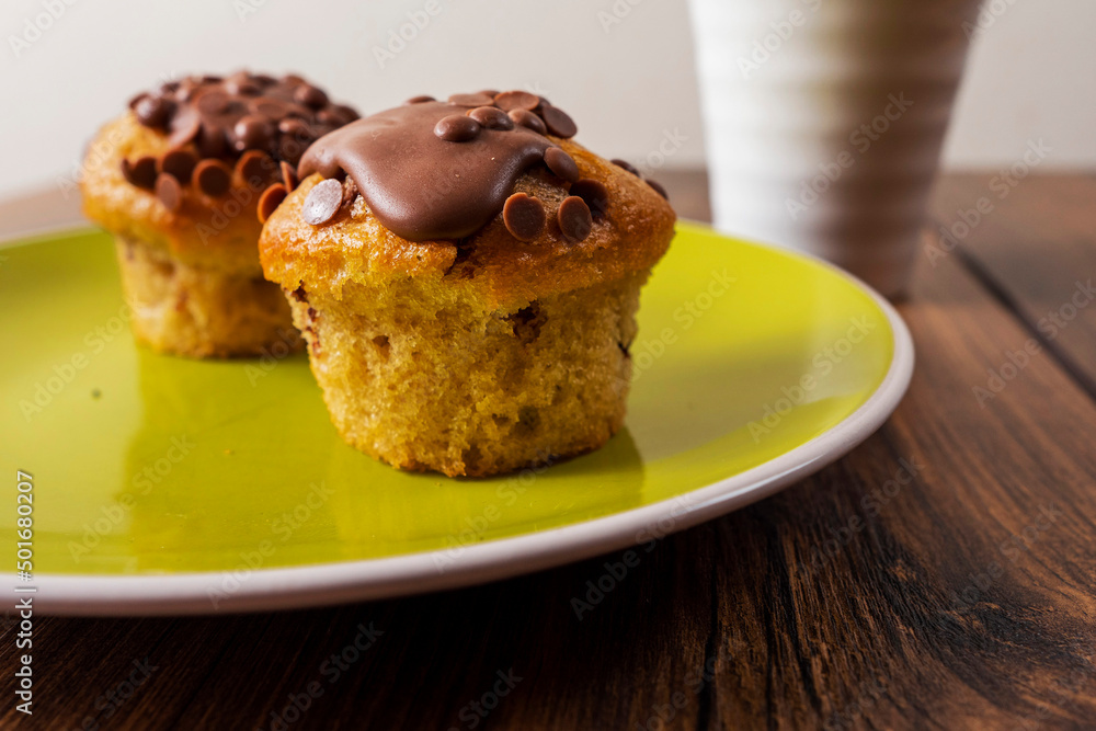 Two chocolate chip muffins on a yellow plate and white cup of tea and wooden table . Copy space. Premium quality bakery product. Two chocolate chip muffins on a yellow plate and white cup of tea and wooden table . Copy space. Premium quality bakery product.