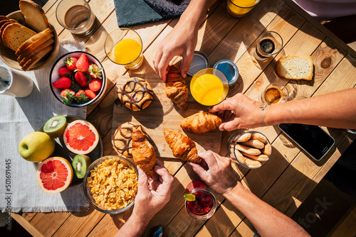 Vertical view of table full of breakfast food and group of people eating and enjoying it using mobile phone. Family and friends eat together and enjoy morning leisure time. Bakery and fruit