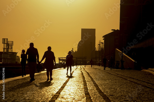Silhouette of people a quay along river Tajus with Monument to the Discoveries in the background in Lisbon against sunset sky