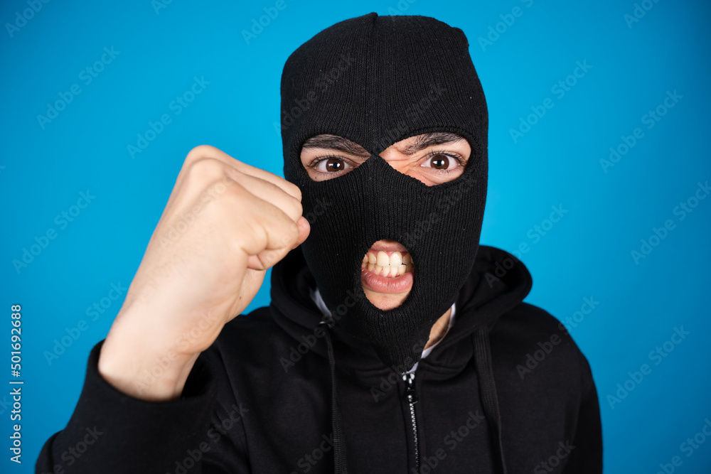 Angry criminal menacing fist up to the camera isolated on blue background. Delinquent wearing face mask.