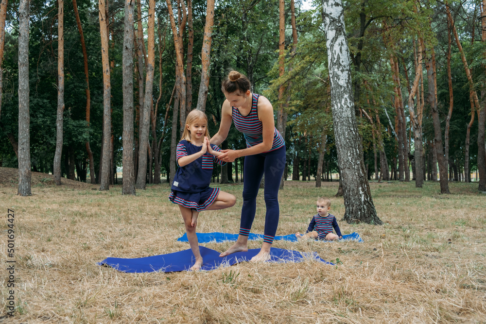 Family fitness and yoga. Happy mom exercising with baby girls on grassy land against trees in forest. Young woman doing yoga exercise outdoors with her daughters together Family fitness and yoga. Happy mom exercising with baby girls on grassy land against trees in forest. Young woman doing yoga exercise outdoors with her daughters together