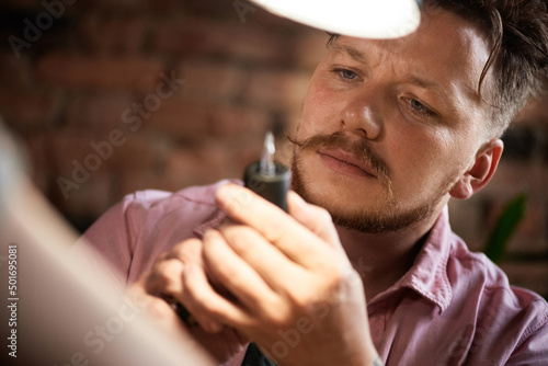 Concentrated male tattooist with beard and moustache looking at tattoo gun attentively under lamp light