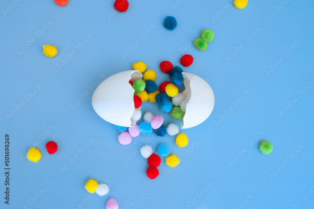 Multi-colored pom-poms falling out of an egg broken into two parts on a blue background, a funny egg
