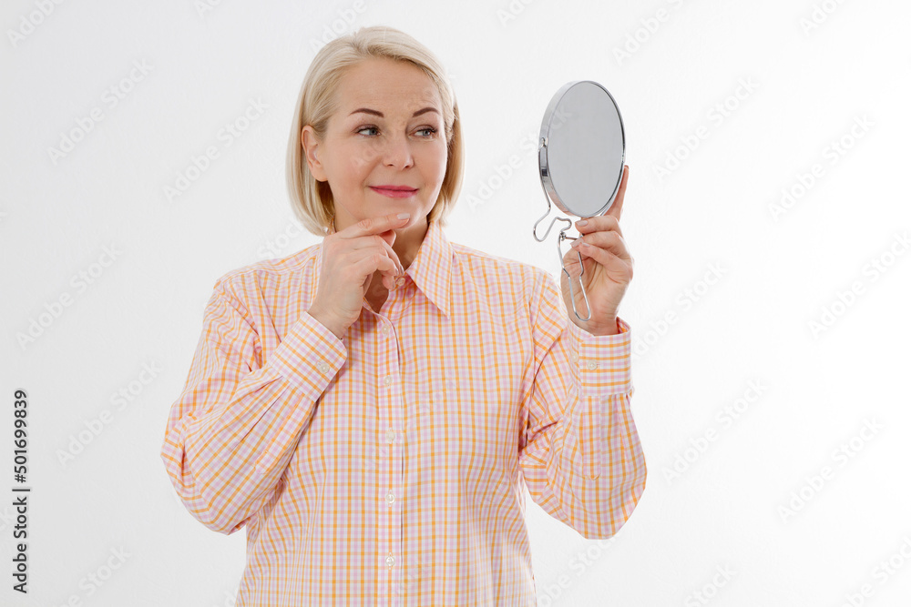 Happy Middle aged Woman looking in mirror on her face with wrinkles on the forehead and wrinkle skin. Collagen and face injections concept. Menopause. Copy space and mock up. Happy Middle aged Woman looking in mirror on her face with wrinkles on the forehead and wrinkle skin. Collagen and face injections concept. Menopause. Copy space and mock up.