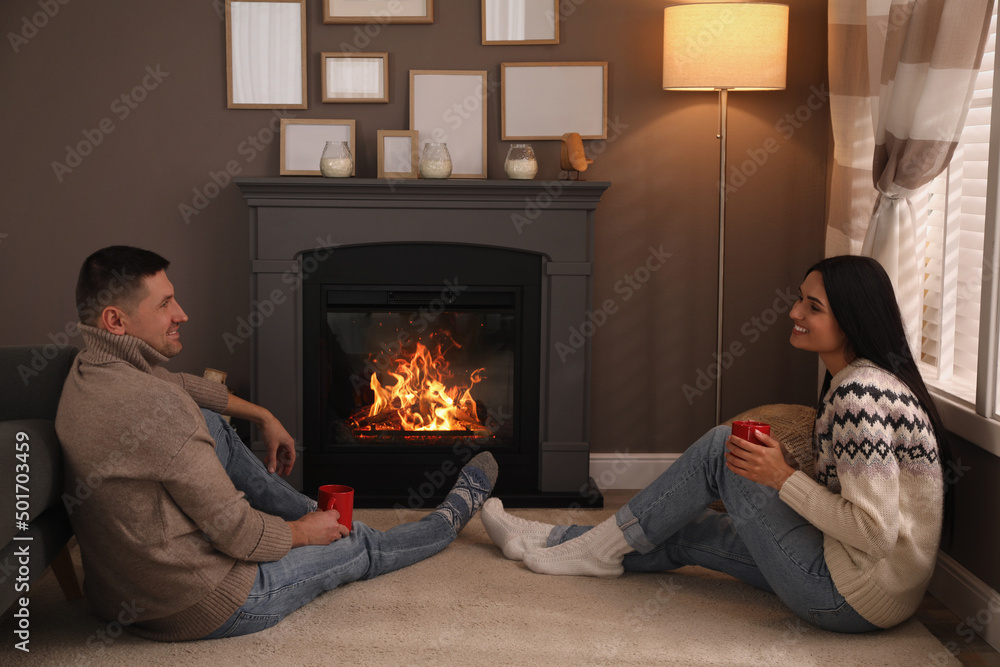 Lovely couple with cups of hot drink spending time together near fireplace at home Lovely couple with cups of hot drink spending time together near fireplace at home