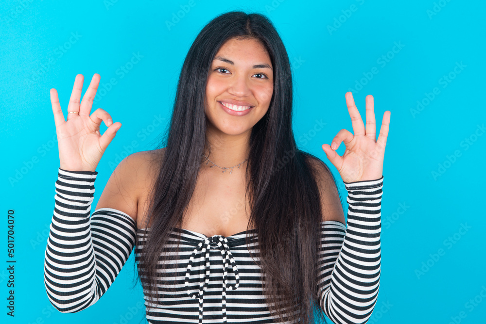 Glad young latin woman wearing striped T-shirt over blue background shows ok sign with both hands as expresses approval, has cheerful expression, being optimistic. Glad young latin woman wearing striped T-shirt over blue background shows ok sign with both hands as expresses approval, has cheerful expression, being optimistic.
