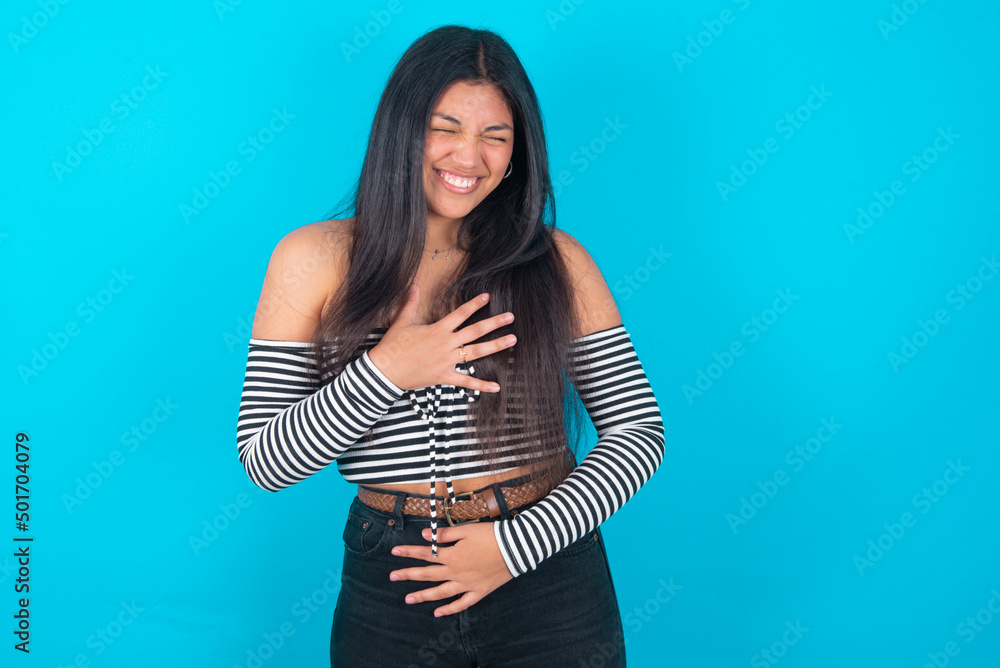 young latin woman wearing striped T-shirt over blue background keeps hands crossed, laughs out loud at good joke, wears casual clothes. young latin woman wearing striped T-shirt over blue background keeps hands crossed, laughs out loud at good joke, wears casual clothes.