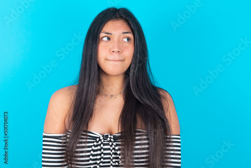 Amazed puzzled young latin woman wearing striped T-shirt over blue background , curves lips and has worried look, sees something awful in front. photo