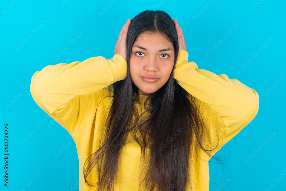 Frustrated young latin woman wearing yellow sweater over blue background plugging ears with hands does not wanting to listen hard rock, noise or loud music.