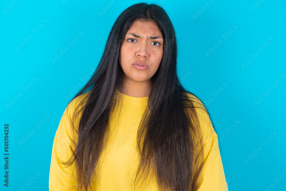 young latin woman wearing yellow sweater over blue background expressing disgust, unwillingness, disregard having tensive look frowning face, looking indignant with something. young latin woman wearing yellow sweater over blue background expressing disgust, unwillingness, disregard having tensive look frowning face, looking indignant with something.