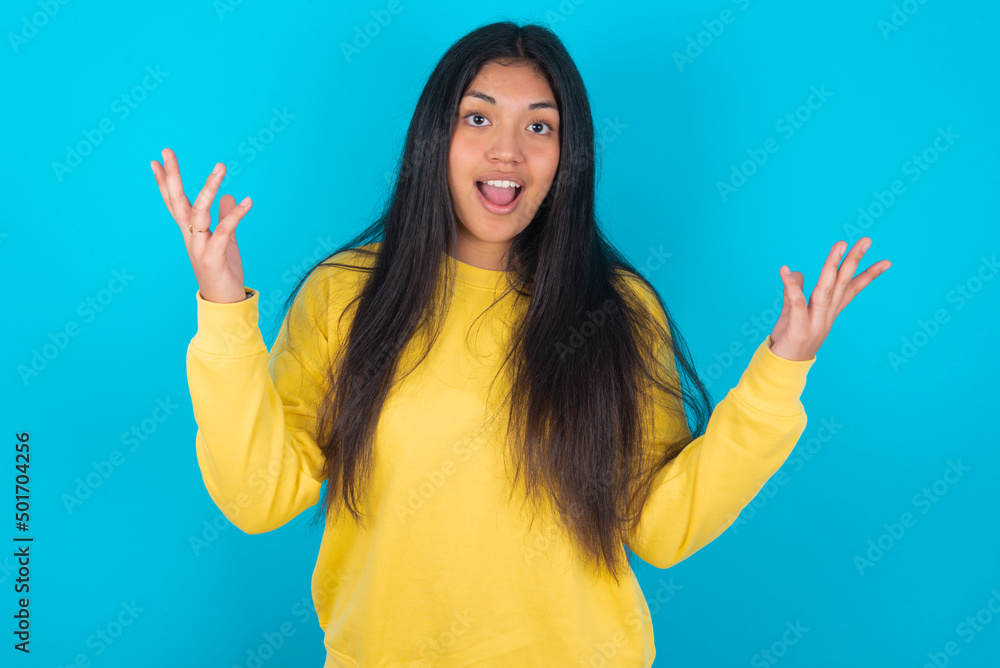 Joyful excited lucky young latin woman wearing yellow sweater over blue background cheering, celebrating success, screaming yes with clenched fists Joyful excited lucky young latin woman wearing yellow sweater over blue background cheering, celebrating success, screaming yes with clenched fists
