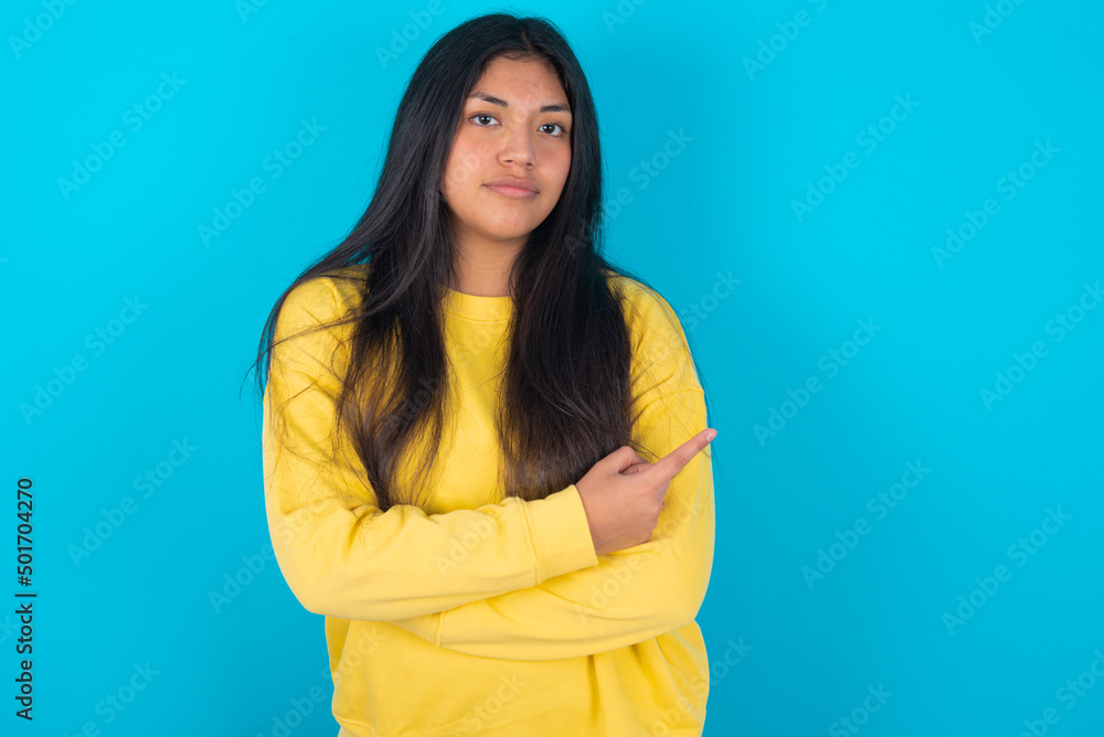 young latin woman wearing yellow sweater over blue background smiling broadly at camera, pointing fingers away, showing something interesting and exciting. young latin woman wearing yellow sweater over blue background smiling broadly at camera, pointing fingers away, showing something interesting and exciting.