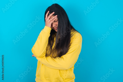 young latin woman wearing yellow sweater over blue background making facepalm gesture while smiling amazed with stupid situation.