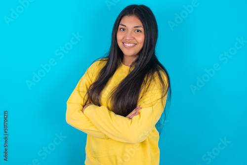 Image of cheerful young latin woman wearing yellow sweater over blue background with arms crossed. Looking and smiling at the camera. Confidence concept.