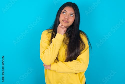 Face expressions and emotions. Thoughtful young latin woman wearing yellow sweater over blue background holding hand under his head, having doubtful look.