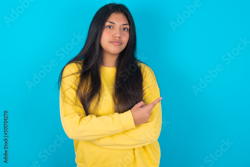 young latin woman wearing yellow sweater over blue background smiling broadly at camera, pointing fingers away, showing something interesting and exciting.