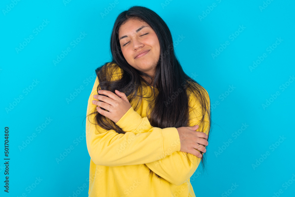 young latin woman wearing yellow sweater over blue background . Hugging oneself happy and positive, smiling confident. Self love and self care. young latin woman wearing yellow sweater over blue background . Hugging oneself happy and positive, smiling confident. Self love and self care.