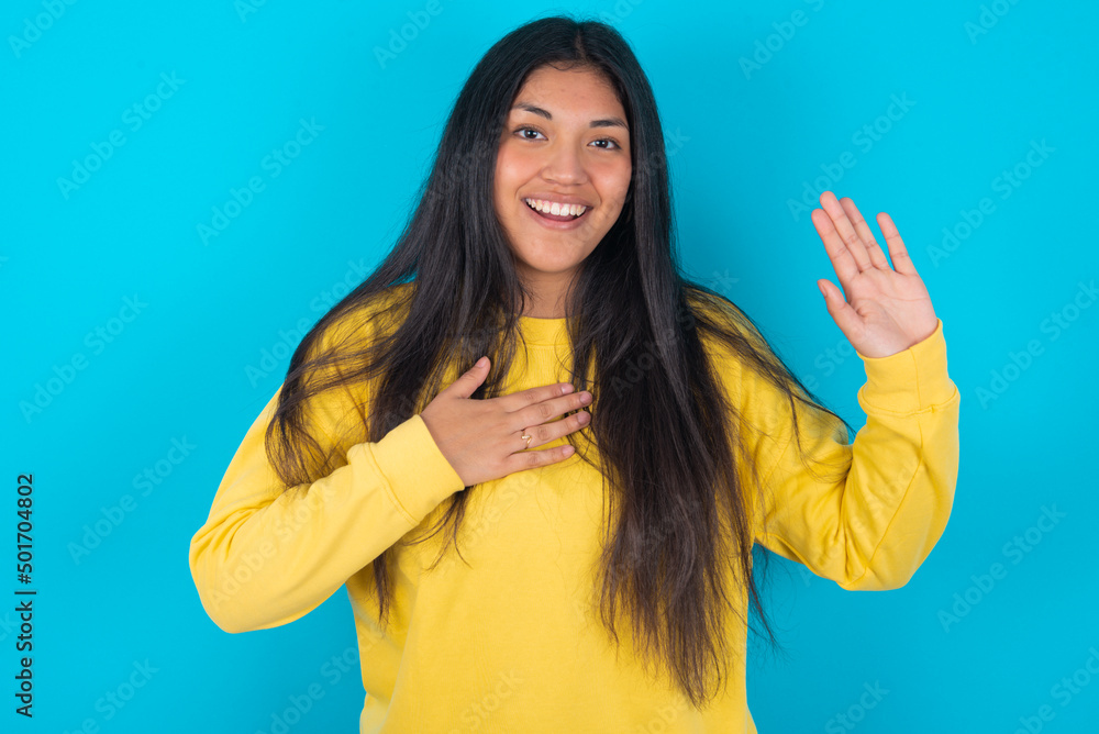 I swear, promise you not regret. Portrait of sincere young latin woman wearing yellow sweater over blue wall raising one arm and hold hand on heart as give oath, telling truth, want you to believe. I swear, promise you not regret. Portrait of sincere young latin woman wearing yellow sweater over blue wall raising one arm and hold hand on heart as give oath, telling truth, want you to believe.