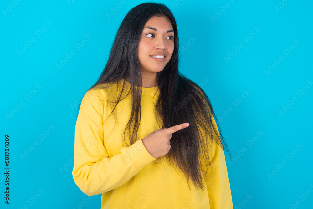 Fototapeta premium Happy cheerful smiling young latin woman wearing yellow sweater over blue background looking and pointing aside with hand. Copy space and advertisement concept.