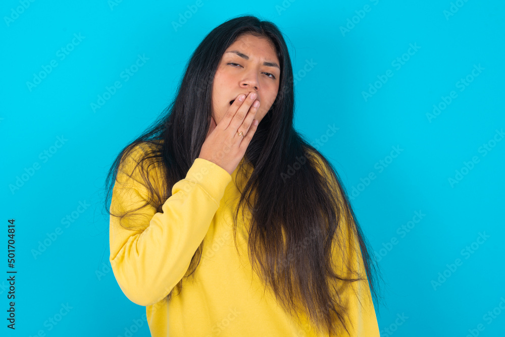 Sleepy young latin woman wearing yellow sweater over blue background yawning with messy hair, feeling tired after sleepless night, yawning, covering mouth with palm. Sleepy young latin woman wearing yellow sweater over blue background yawning with messy hair, feeling tired after sleepless night, yawning, covering mouth with palm.