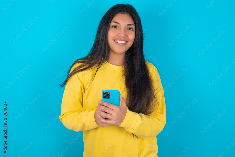 Smiling young latin woman wearing yellow sweater over blue background friendly and happily holding mobile phone taking selfie in mirror. Smiling young latin woman wearing yellow sweater over blue background friendly and happily holding mobile phone taking selfie in mirror.