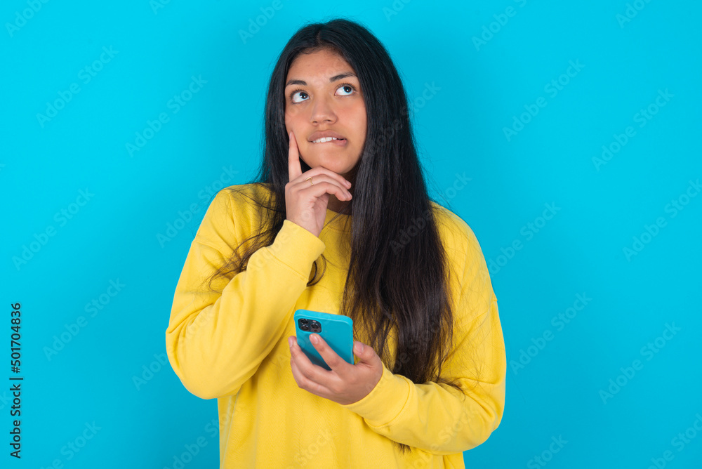 Image of a thinking dreaming young latin woman wearing yellow sweater over blue background using mobile phone and holding hand on face. Taking decisions and social media concept. Image of a thinking dreaming young latin woman wearing yellow sweater over blue background using mobile phone and holding hand on face. Taking decisions and social media concept.