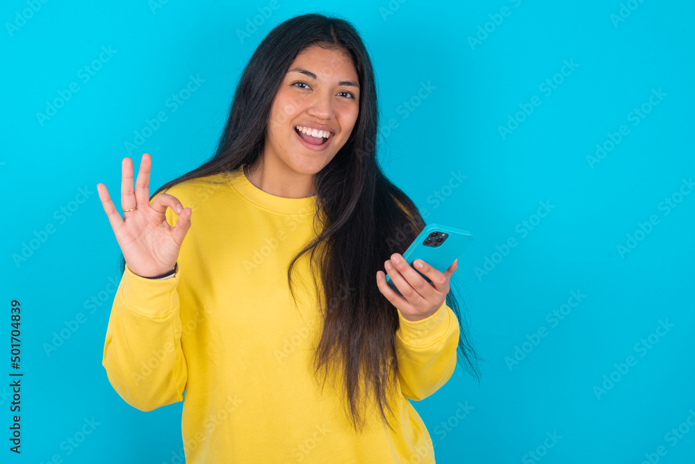 Happy young latin woman wearing yellow sweater over blue background sending a message on his smartphone or taking a selfie and making ok sign with his hand. Happy young latin woman wearing yellow sweater over blue background sending a message on his smartphone or taking a selfie and making ok sign with his hand.