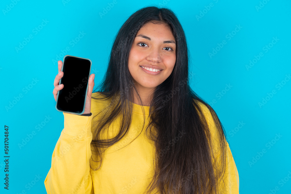 Smiling young latin woman wearing yellow sweater over blue background showing empty phone screen. Advertisement and communication concept. Smiling young latin woman wearing yellow sweater over blue background showing empty phone screen. Advertisement and communication concept.