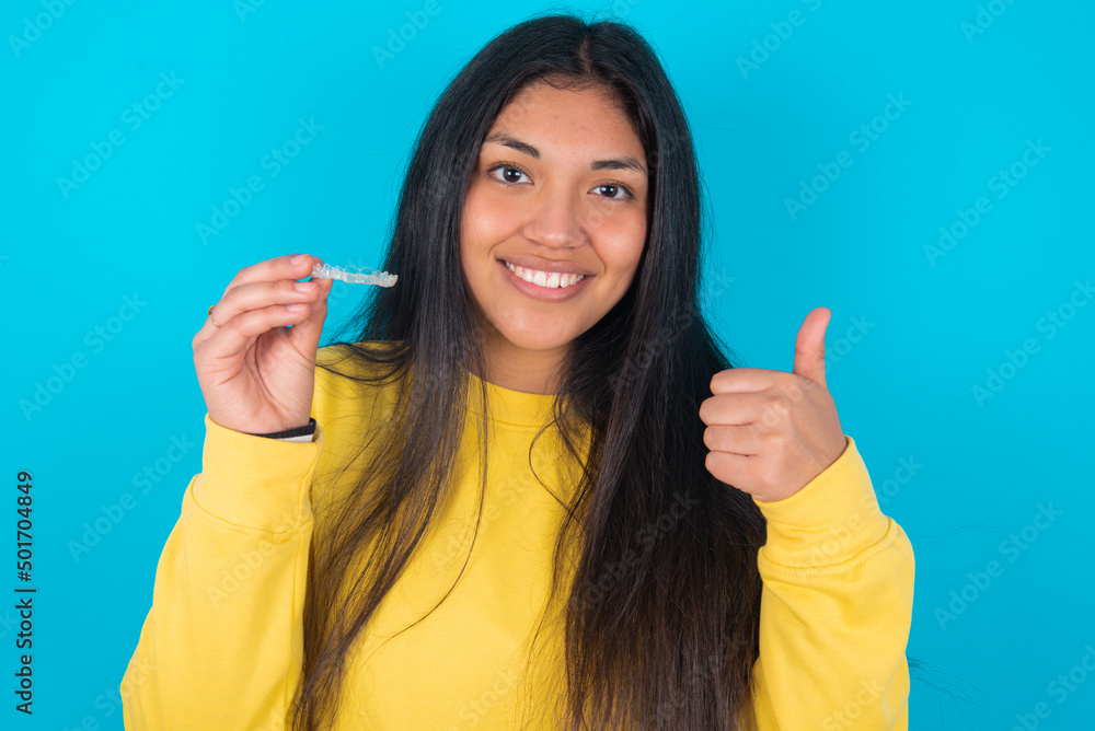 young latin woman wearing yellow sweater over blue background holding an invisible braces aligner and rising thumb up, recommending this new treatment. Dental healthcare concept.