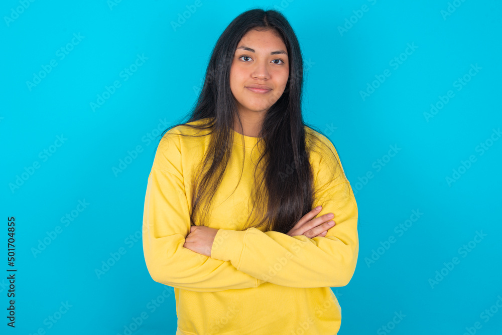 Self confident serious calm young latin woman wearing yellow sweater over blue background stands with arms folded. Shows professional vibe stands in assertive pose. Self confident serious calm young latin woman wearing yellow sweater over blue background stands with arms folded. Shows professional vibe stands in assertive pose.